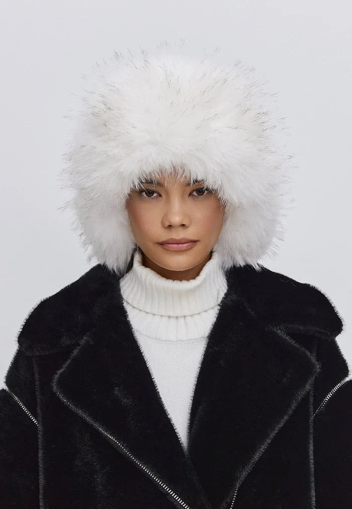 Woman wearing a stylish winter fashion hat, the Irina faux fur hat in black and white, paired with a black coat and white turtleneck.