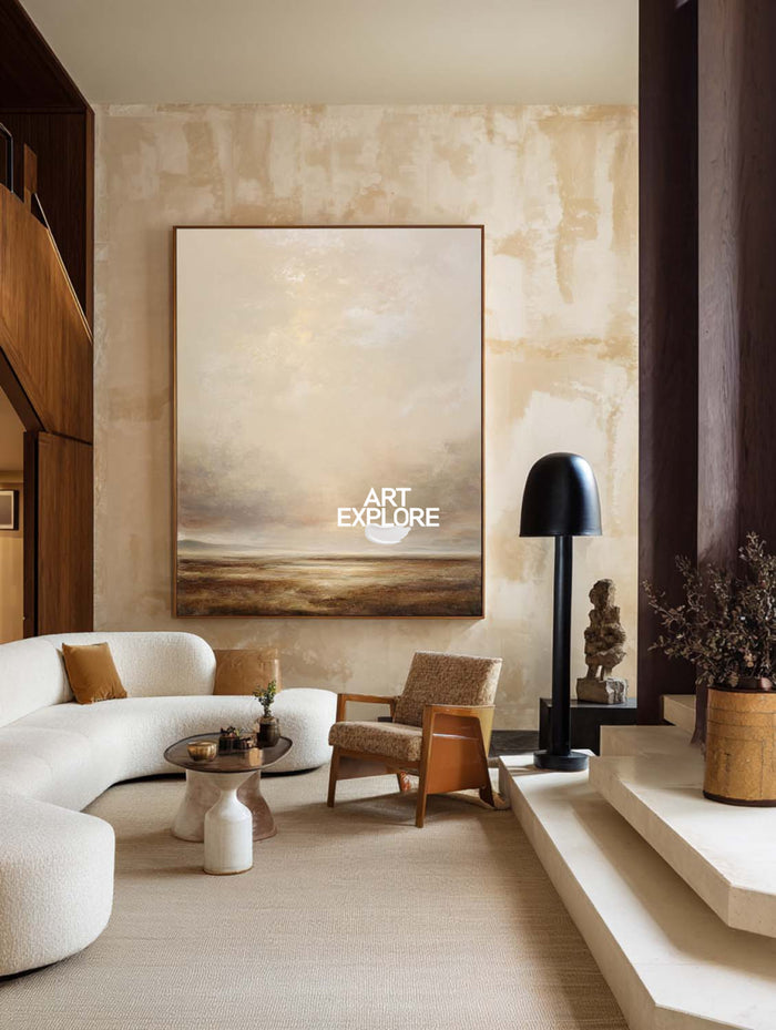 A large vertical landscape painting of a beach or sea under a cloudy sky, displayed on a textured beige wall in a modern, warm-toned living room with a curved white sofa, wooden chair, and black floor lamp.